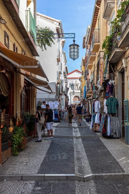 Lively shopping street with people between historic buildings under a blue sky, Granada
