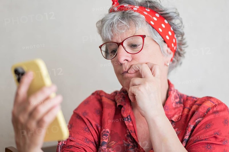 A senior woman wearing glasses and a red headband is holding and looking at a smartphone, contemplating something with a thoughtful expression, dressed in a red floral shirt