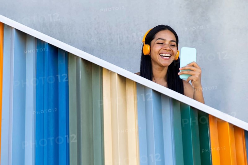 A joyful woman with headphones smiles while using her smartphone against a backdrop of vibrant modern architecture, conveying a sense of connection and technology