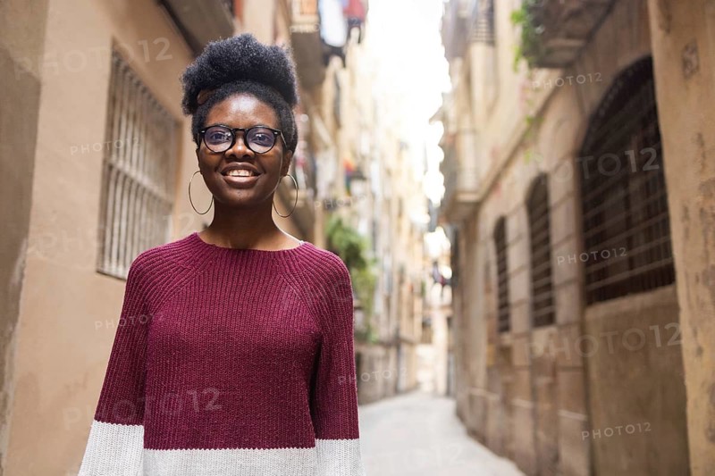 A cheerful woman wearing a burgundy sweater and glasses smiles confidently while standing in a charming, narrow urban alleyway framed by rustic buildings and windows
