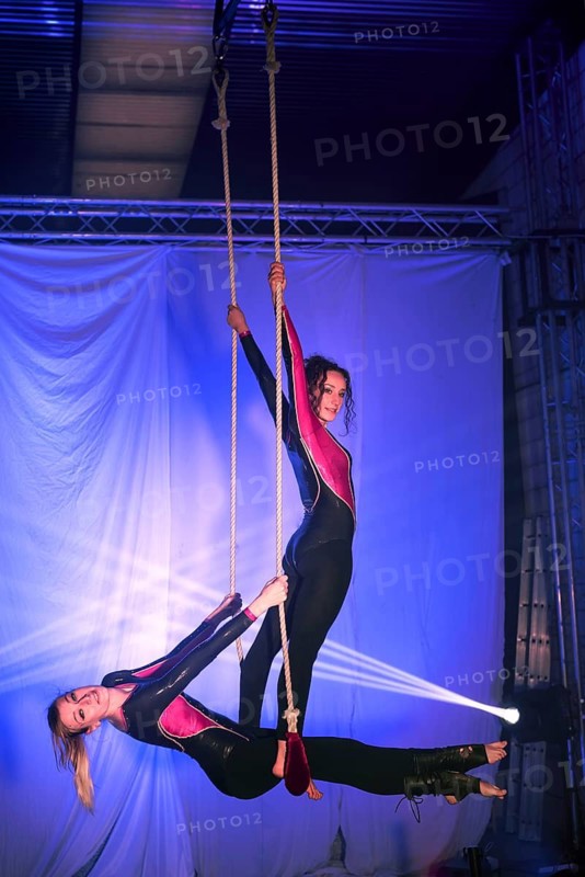 Two trapeze artists performers execute an elegant aerial routine on ropes, dressed in shiny black and pink outfits, with a vivid blue and purple lit stage backdrop enhancing the scene