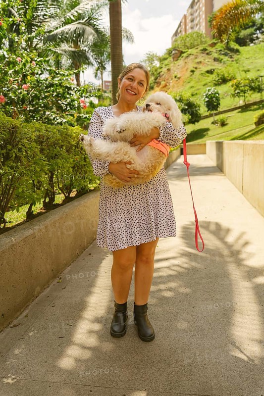 Smiling woman holding her small white dog while standing on a paved path in a park, surrounded by greenery and buildings in the background, enjoying a sunny day outdoors