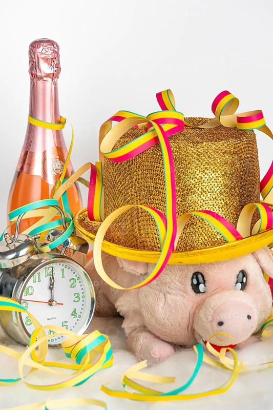 Plush pig wearing a golden hat next to an alarm clock and champagne bottle, colourful streamers, New Year's Eve