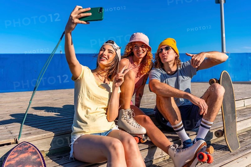 Three young, stylish friends are sitting on a wooden boardwalk, taking a selfie with a smartphone while wearing roller skates and enjoying the sunny weather