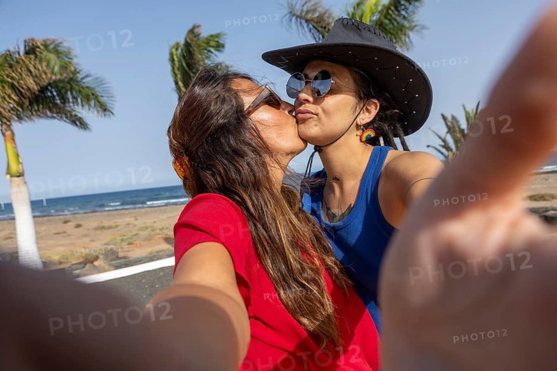 A joyful lesbian couple shares a tender moment by the beach, capturing a selfie on a sunny summer day. The scene exudes love, happiness, and LGBTQIA+ pride