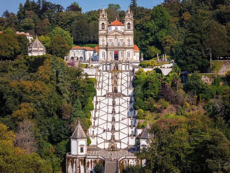 Drone aerial view of the Sanctuary of Bom Jesus do Monte in Braga, Portugal, featuring the church, grand staircase, gardens, and forested hillside in sunny daylight