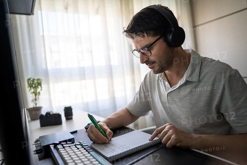 A man wearing headphones is focused on studying Mandarin online at home. He is taking notes on a notepad while sitting at a desk with natural light streaming in