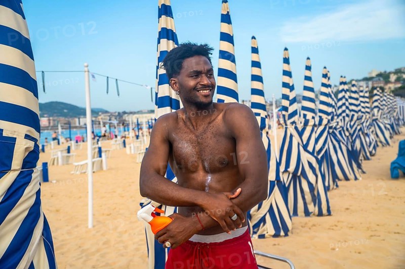 Young shirtless man standing on a sandy beach, joyfully applying sunscreen to his arm, with rows of blue and white striped parasols in the background on a sunny day
