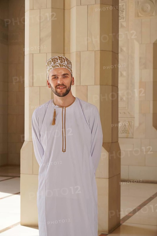 A young male wearing a traditional white dishdasha and embroidered kumma stands inside Sultan Qaboos Mosque, Muscat, Oman. His relaxed and friendly demeanor complements the elegant, sunlit backdrop of the mosque's columned hall