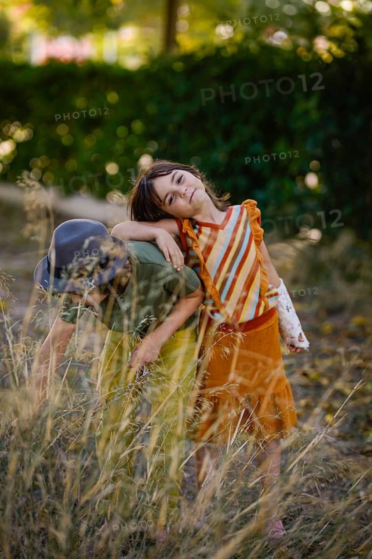 Two children enjoy a sunny day in a meadow, surrounded by tall grass and lush greenery. The moment captures carefree childhood and nature's beauty in warm, natural light