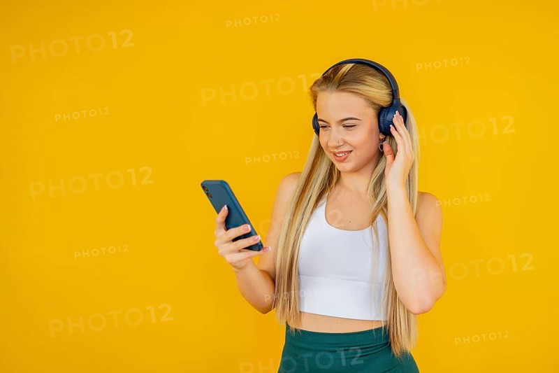 A young woman in a white top and green pants enjoys music through headphones while holding a smartphone. She stands against a vibrant yellow background at home, expressing joy