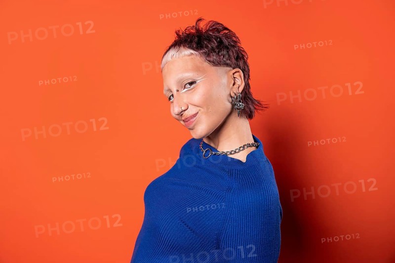 Fashion-forward woman with an edgy hairstyle and bold makeup wearing a vibrant blue outfit against an orange background, showcasing unique earrings and a chain necklace