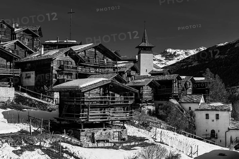 Snow-covered roofs of old farmhouses and barn storage, in the back the large wooden cross and the church tower of the Saint Theodule church, black and white photo, Grimentz, Val d'Anniviers, Valais Alps, Canton of Valais, Switzerland