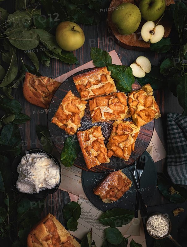 Sliced apple pie on wooden board, apples, leaves, whipped cream, from above, food photography