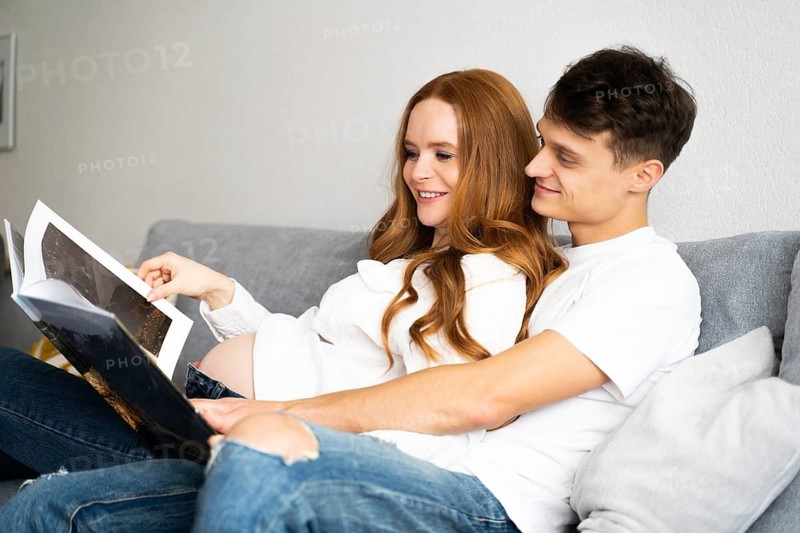 A young couple sits on the couch, joyfully anticipating the arrival of their baby as they look at a photo album, capturing a tender moment of love and excitement for the future