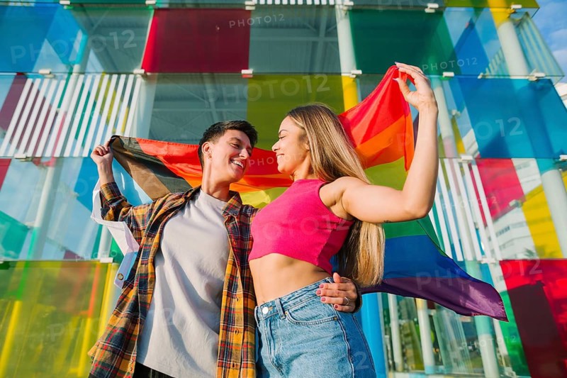 A joyful lesbian couple embraces while holding the Pride and Transgender flags. Their loving gaze and vibrant background symbolize inclusivity, self-expression, and the celebration of diversity