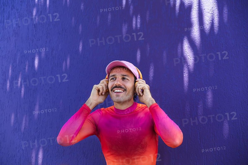 Cheerful young Spanish man with a mustache is captured in Quito, Ecuador. He wears vibrant pink clothing and enjoys listening to music on headphones, reflecting a joyful urban lifestyle and self-expression themes relevant to the LGTBIQ+ community