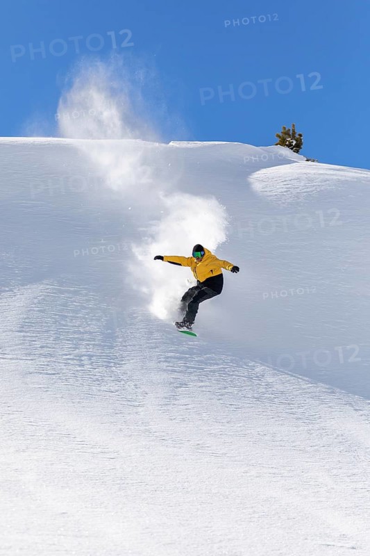 A snowboarder in a yellow jacket glides effortlessly down a snowy slope, kicking up powder under a clear blue sky. The scene captures the thrill and freedom of snowboarding