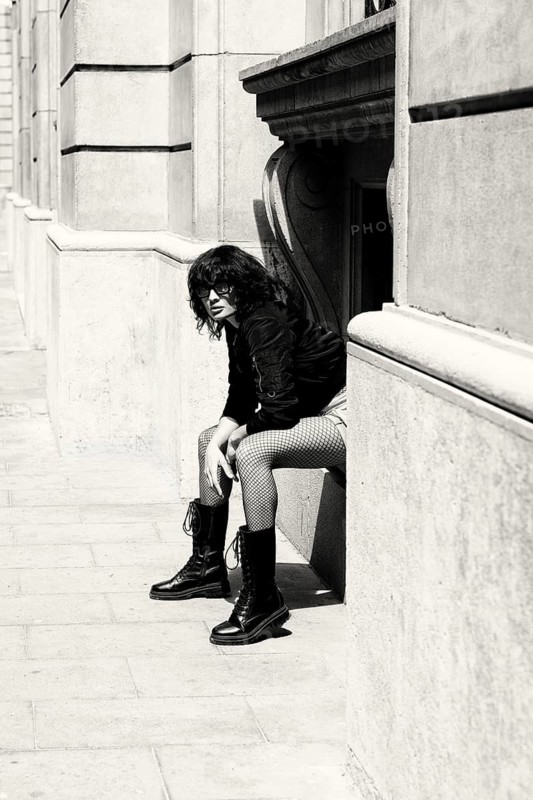A fashionable woman wearing bold boots and patterned pants sits against a historic building. Captured in black and white, the high-contrast photo exudes attitude, confidence, and urban style