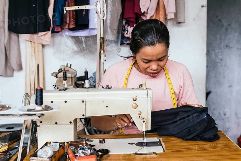 A asian seamstress focuses on sewing in a small workshop. Surrounded by tools and garments, she uses a vintage sewing machine, embodying craftsmanship and tradition
