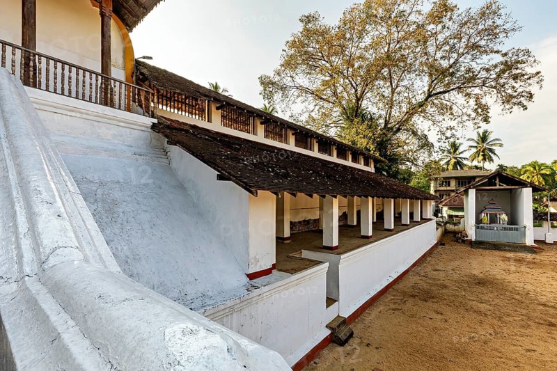 White building structure with wooden railings and tropical vegetation in the background at sunset, The Sri Sumana Saman Devalaya Temple near Ratnapura in Sri Lanka