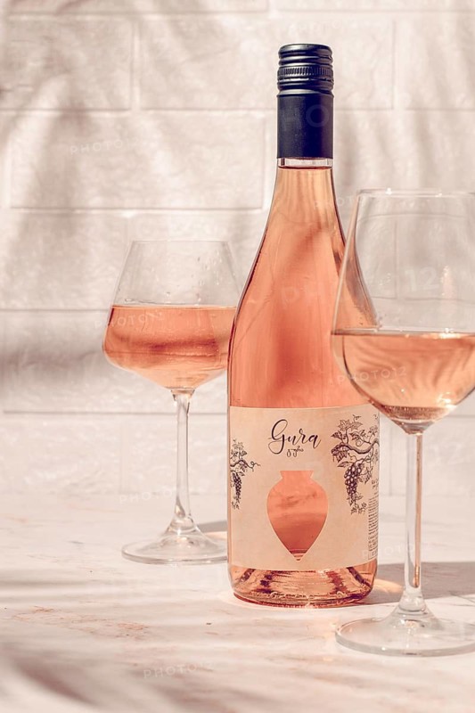 A bottle of Gura wine sits on a table beside two glasses filled with wine. The bright setting highlights the wine's color. This scene shows a moment of enjoyment and relaxation
