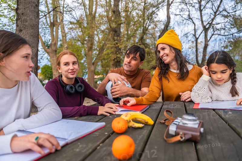 Family members sitting at a wooden picnic table, spending quality time together, collaborating on homework and learning outdoors, surrounded by nature and autumn trees