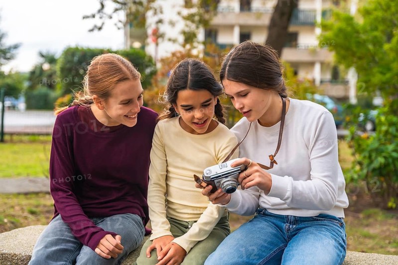 Three friends sitting on a park bench smiling as they review photos on a vintage camera, sharing a joyful, relaxed moment of connection and outdoor leisure together