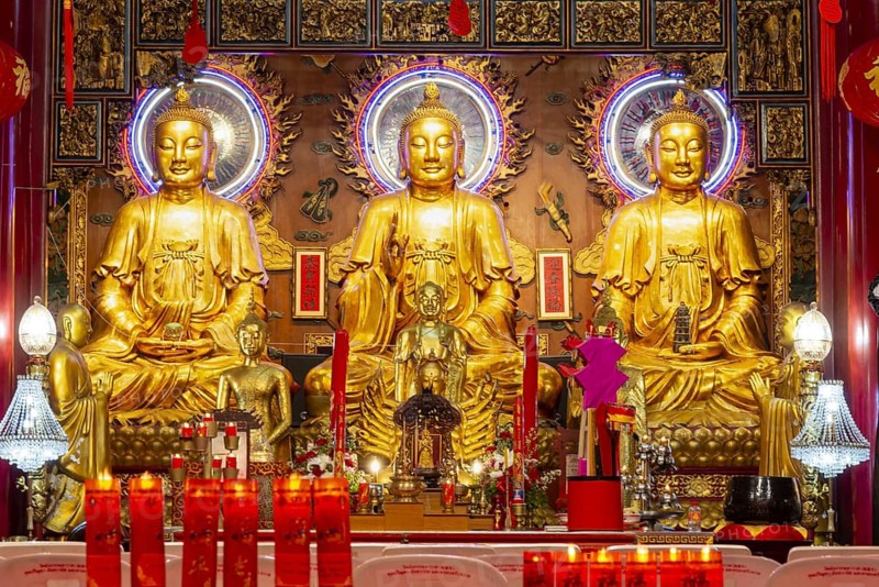 A quiet moment in the heart of a Chinese-Buddhist temple: Three golden Buddha figures shine in warm light, surrounded by filigree ornaments, red inscriptions and offerings that fill the room with an atmosphere of devotion and timeless dignity. The composition of light, gold and ritual objects condenses into a place of inner collection where wisdom, compassion and spiritual presence can be felt. Wat Mangkon Kamalawat, Yaowarat Road, Chinatown, Bangkok, Tailandia