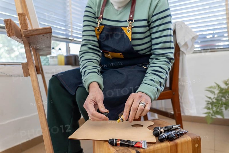 Close-up of a elderly artist's hands squeezing oil paint from a tube onto a wooden artist palette, with other paint tubes laid out on a small studio stool