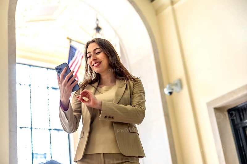 Businesswoman in a brown suit smiling as she scrolls a smartphone inside a bright, arched manhattan building with an american flag, conveying professional connection and confidence