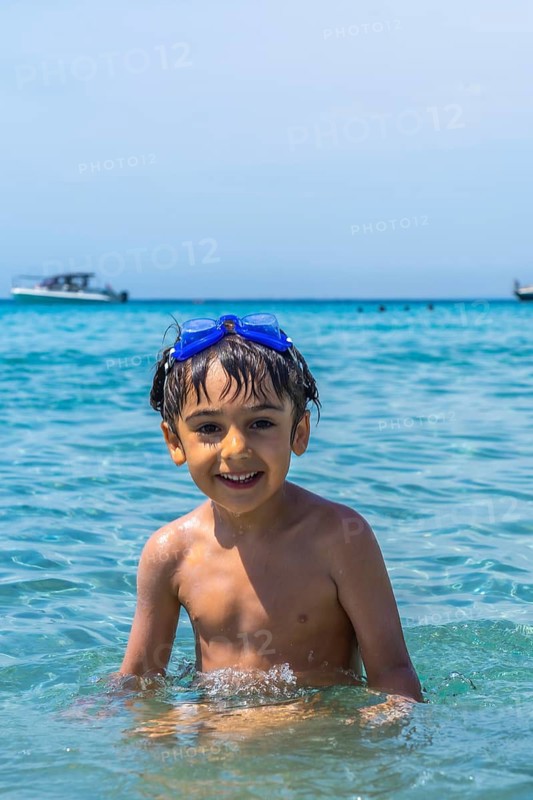 Happy little boy wearing swimming goggles, smiling directly at the camera while standing in the clear blue sea during a summer holiday on tabarca island, alicante, spain