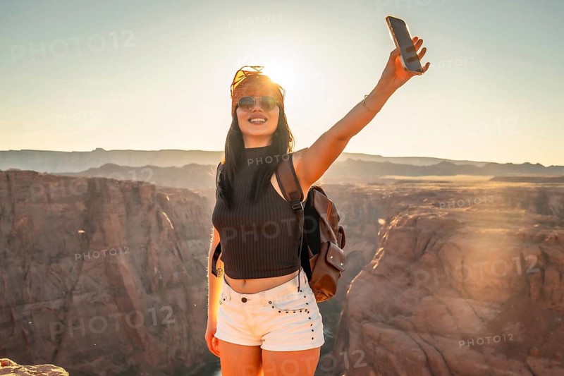 Young woman smiling and holding a smartphone, capturing a selfie against the vast desert canyon landscape and glowing sun during a vibrant orange and yellow sunset