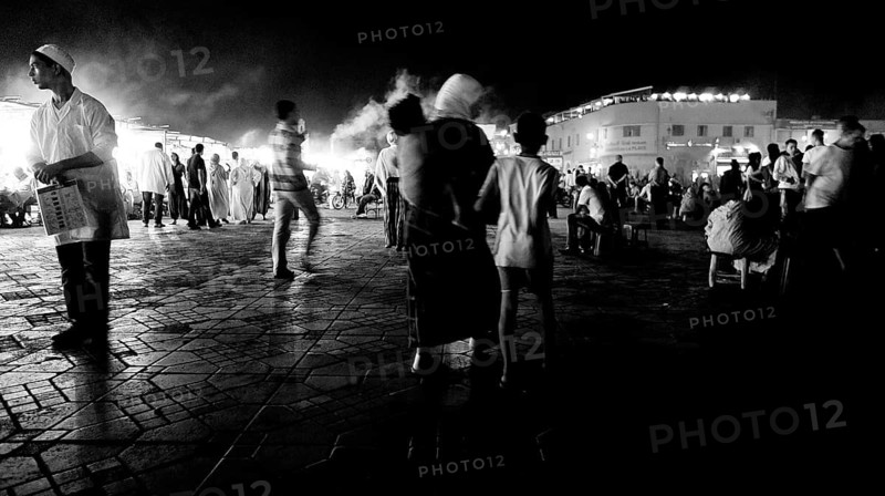 Groups of people strolling on a paved black and white night market, evening food stalls at Djemaa el Fna, Marrakech, Morocco