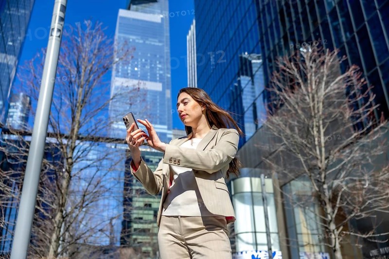 Businesswoman looking at her mobile phone, navigating or scrolling content outdoors in a modern urban financial district with skyscrapers forming the backdrop