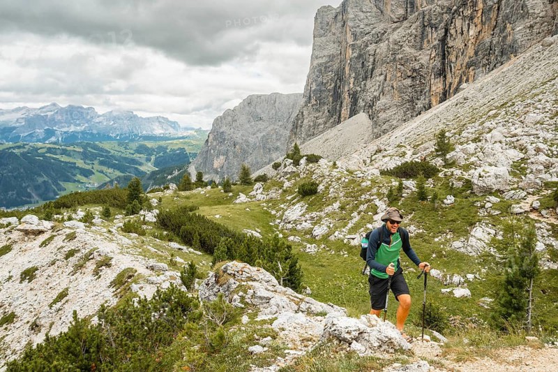 Active hiker with backpack and trekking poles walking rocky path through green alpine pass steep limestone massifs background Puez-Odle Dolomites Alps Italy summer landscape