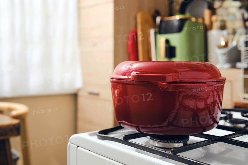 Red cast iron pot rests on stove in cozy kitchen. Wooden furniture and soft light from the window create inviting space. Tools and appliances are visible in background