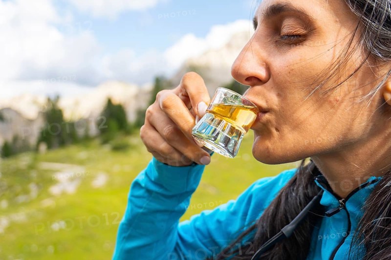Close up woman traveler drinking local herbal spirit schnapps or grappa shot glass wooden terrace green mountain valley backdrop Dolomites Italy. Fragrant, grape-based pomace home made brandy