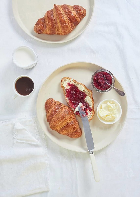 Croissants with butter and jam for continental breakfast coffee and milk on white tablecloth table, directly above