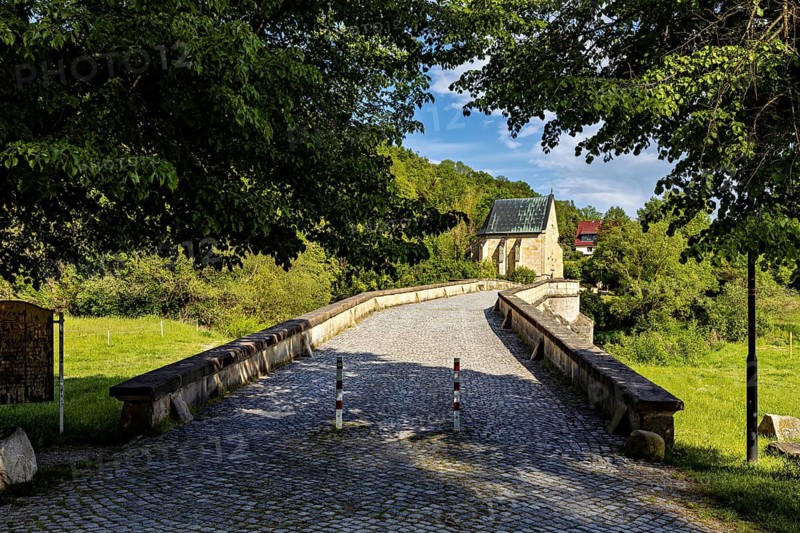 An old stone bridge leads across a quiet river to a small church in the countryside on a sunny day, The Liborius Chapel on the Werrbrücke near Creuzburg in Thuringia