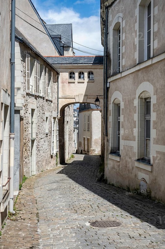 Narrow, sunlit alley with historic cobblestones and historic stone buildings, dominated by an architectural archway superstructure with windows in the old town of Angers, Maine-et-Loire, Pays de la Loire, France
