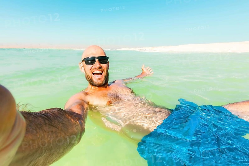 POV caucasian male Man floating wave pose send greetings in turquoise Dead Sea water wearing a traditional keffiyeh and sunglasses. Relaxing summer vacation at the lowest point on Earth in Jordan
