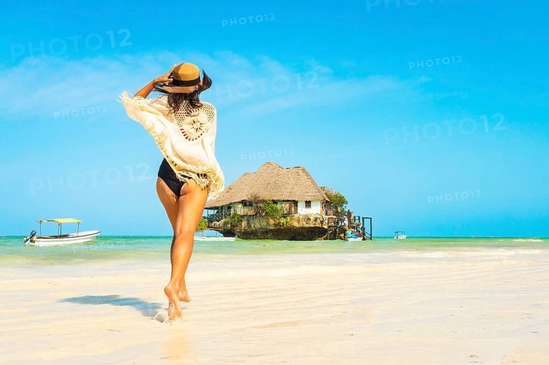Excited one traveler woman in black bikini runs through shallow turquoise water beach to iconic Rock Restaurant in Zanzibar. High-energy dynamic shot capturing pure joy and summer freedom in paradise