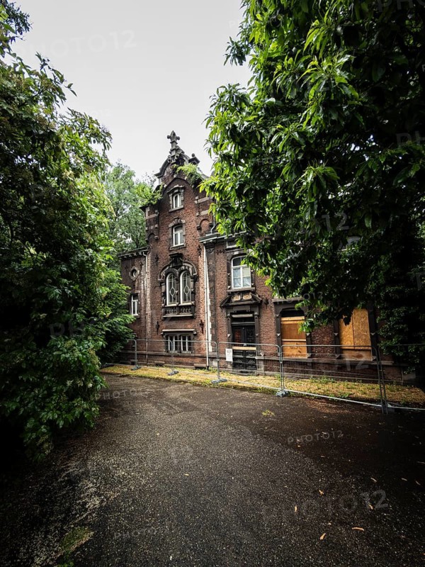 An old brick villa stands abandoned and boarded up with wooden panels behind a construction fence, surrounded by thick, green trees, Liège, Belgium