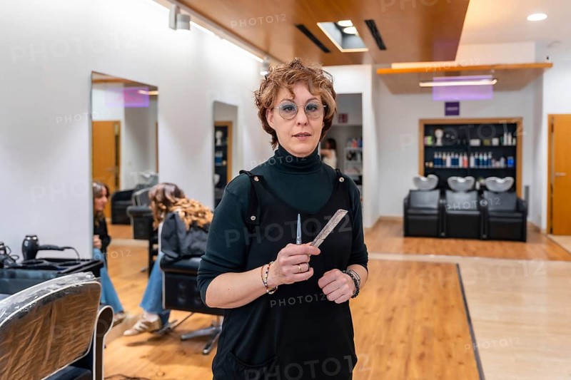 Professional female hairstylist standing confidently with short curly hair and glasses, holding hairdressing scissors and a comb in a modern beauty salon setting