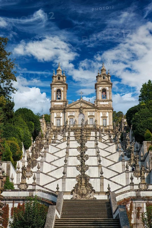 Vertical view of Bom Jesus do Monte in Braga, Portugal, highlighting the upper staircase, baroque statuary and twin bell towers framed by dramatic clouds over the sanctuary