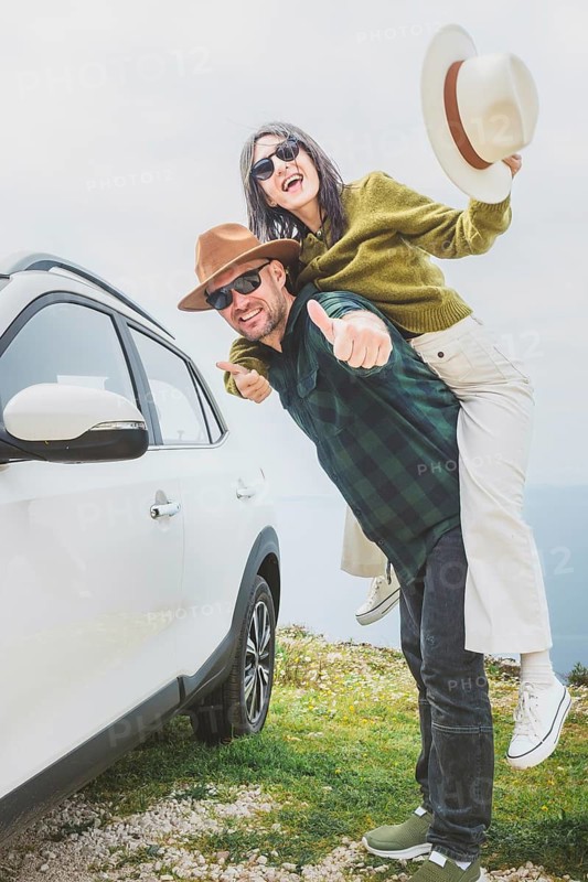 Portrait Happy couple Man with woman having fun piggyback next to white car on coastal cliff. Vast blue ocean and mountain horizon create romantic, joyful travel mood. Summer holiday adventure vibes
