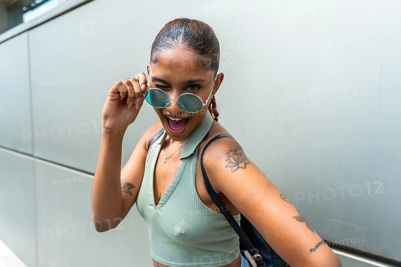 Young woman showing playful expression and winking, pulling down her sunglasses while happily posing outdoors, displaying her unique personality and tattoos