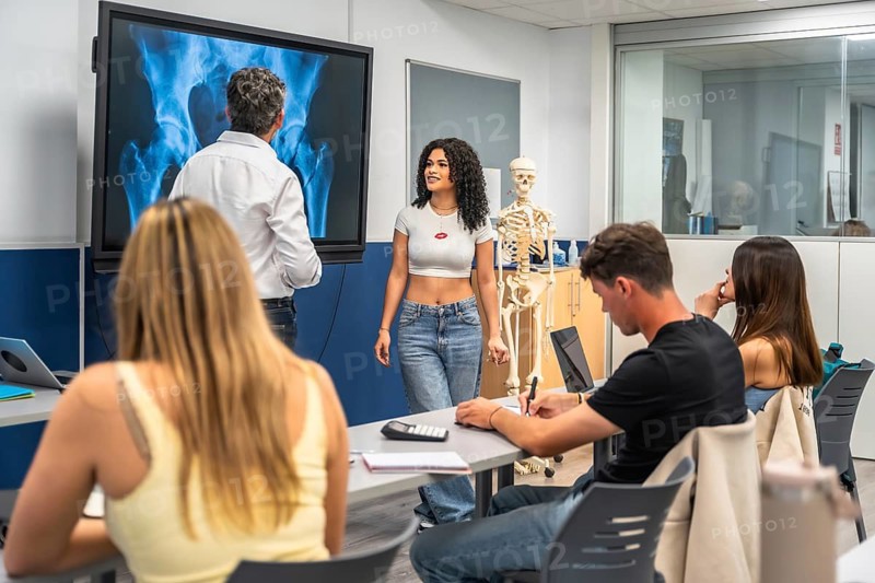Professor explaining human anatomy to university students using an x ray image of a pelvic bone on a digital screen, with a skeleton model present in the classroom during a medical education lesson
