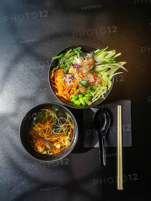 Japanese-style lunch. Bowl with fresh salmon, carrots, cucumbers, green beans, chukka lettuce. Shiro miso soup. Wooden chopsticks and Asian spoon on black table at Asian restaurant. Neatly arranged meal with two bowls placed side by side on dark surface. One bowl contains clear noodle soup with vegetables, while other features colorful poke bowl with salmon, edamame, carrots, cucumber, seaweed and sesame seeds. A black spoon on a napkin and a pair of chopsticks complete the modern healthy food presentation